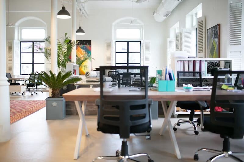 Interno di un ufficio moderno con scrivanie eleganti, sedie ergonomiche e ampie finestre che offrono abbondante luce naturale. Lo spazio presenta un design contemporaneo con linee pulite, mobili minimalisti e un'atmosfera professionale e accogliente. La combinazione di colori è neutra, con tonalità di grigio, bianco e accenti in legno, creando un ambiente di lavoro produttivo e raffinato.