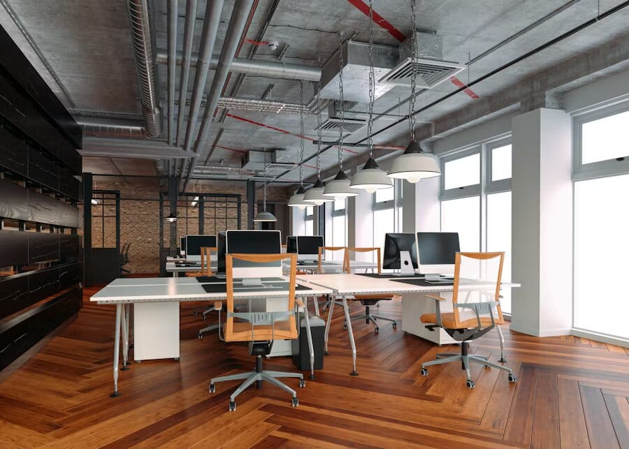 Ufficio moderno open space con tubature a vista, lampade sospese e pavimento in legno, dotato di computer su scrivanie bianche ordinate.