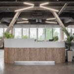 Vista frontale di una reception moderna con bancone in legno e bianco, circondata da piante e grandi finestre in un ufficio open space industriale.