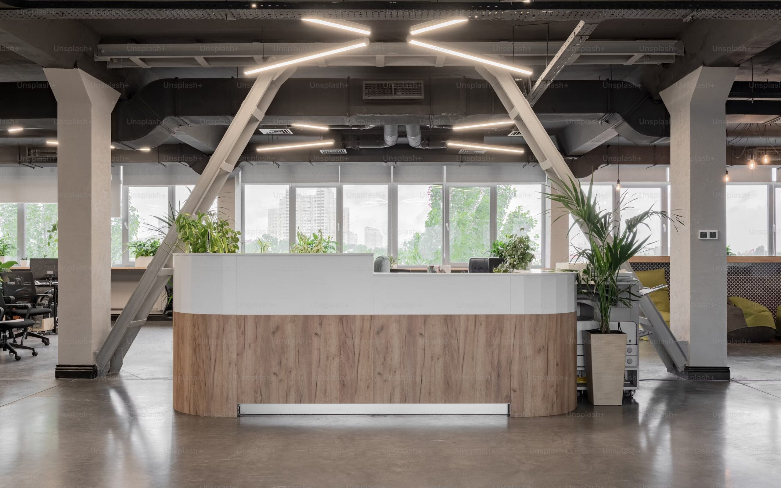 Vista frontale di una reception moderna con bancone in legno e bianco, circondata da piante e grandi finestre in un ufficio open space industriale.